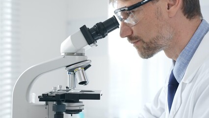 Adult man scientific researcher analyzing sample through microscope, wearing protective gear in high tech laboratory environment. Medicine and science concept