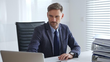 Focused businessman sitting at a modern desk, working intently on a laptop surrounded by folders,...