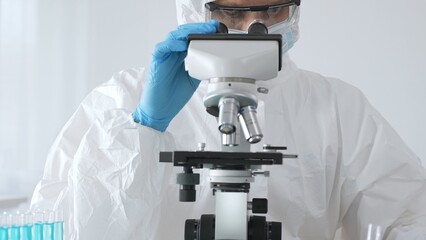 A male doctor in a protective suit and gloves uses a microscope while analyzing samples, conducting...
