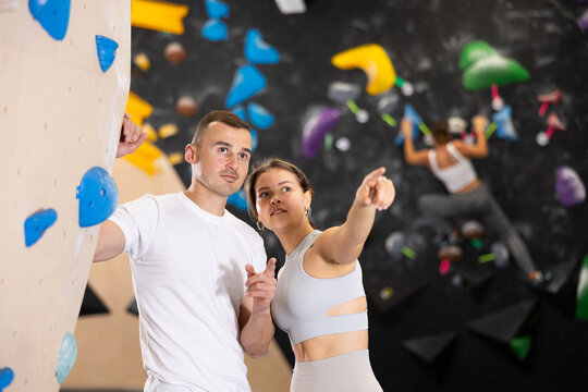 Interested young girl and man standing near bouldering wall in climbing gym, engaged in conversation. Coach instructing beginner female climber about specific moves or technique