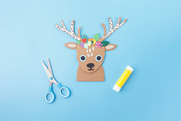 Handmade reindeer head with floral crown and crafting supplies on blue surface for kids art classes, stationery design, festive DIY sets, hobby retail packaging, and seasonal craft businesses,