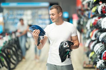 Young man carefully examining and comparing two different bike helmets, considering style and safety features before making purchase in cycling equipment store