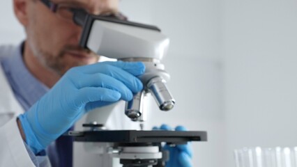 Close up of male scientist in white coat and blue gloves adjusting microscope, conducting pharmaceutical research in modern laboratory. Medicine concept