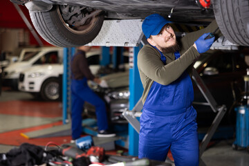 Skilled male auto mechanic in blue overalls repairing automobile in car workshop