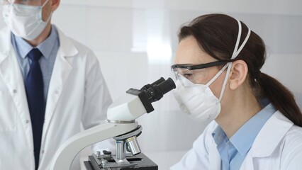 Two scientists in lab coats and protective gear conduct research in a sterile laboratory: one observes through a microscope, and the other holds a test tube with turquoise liquid. Medicine and science