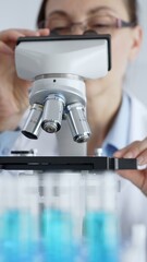Female scientist wearing protective equipment checking microscope, analyzing blue test tubes in modern laboratory research setting. Medicine, healthcare and science concept