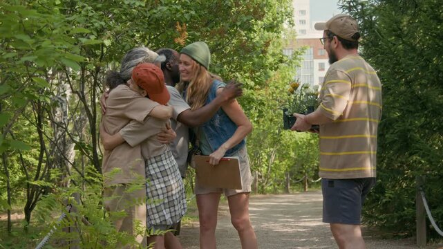 Full shot of happy Caucasian mother, father and young son walking down alley of local park carrying tray of flowers, meeting multiethnic neighbors, greeting and hugging while preparing to plant plugs