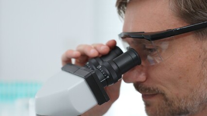 Mature scientist examining specimen under microscope, wearing protective eyewear in contemporary...
