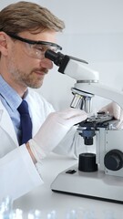 Researcher examining sample through microscope, wearing safety eyewear in sterile laboratory setting, surrounded by scientific glassware. Medicine, healthcare and science concept