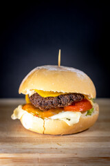 Close-up of a double beef cheeseburger with cheddar and mozzarella cheese, tomato slices, lettuce, and sauce in a fresh bun, presented on a wooden cutting board against a dark background.