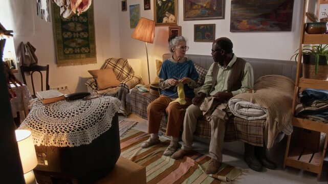 High angle shot of elderly interracial couple sitting on couch and chatting while knitting and watching retro TV in their small cozy living room