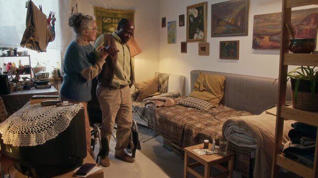 Loving elderly woman helping her African American husband with walking disability get from wheelchair to couch in small poor living room