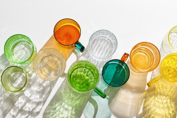 Summer still life scene with colored glass cups. Glittering glasses for water or tea on white table background at sunlight. Long harsh shadows and caustics. Summer vacation concept, top view