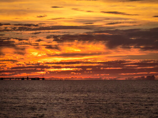 Striking view of a fiery orange and red sunset over the ocean, with a distant pier silhouette beneath dramatic cloud formations.
