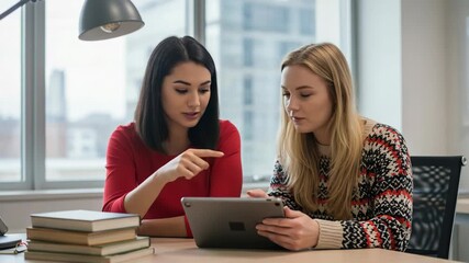 Two young female students collaborating on a digital tablet while studying together at a desk with books in a modern learning environment - Powered by Adobe