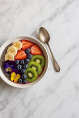 Vibrant smoothie bowl topped with fresh kiwi, strawberries, blueberries, banana slices, chia seeds, and edible flowers, served with a vintage spoon on marble.