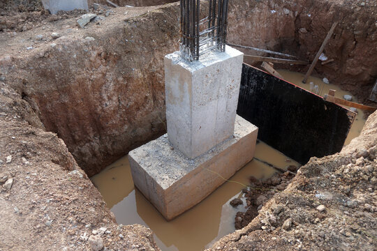 A large rectangular concrete foundation and piers protruding from the foundation are in a hole filled with standing water.