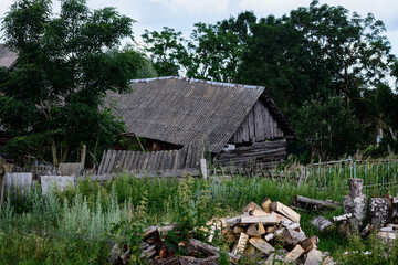 Old wooden house surrounded by overgrown grass and stacked firewood in a rural setting