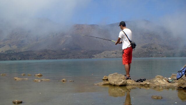 Man fishing on the rocky shore of a crater lake with misty mountains in the background, perfect for nature, leisure, and travel themes