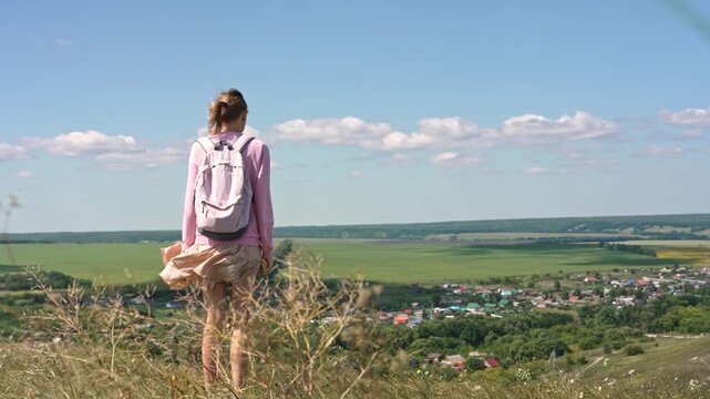 A woman stands on a hilltop, taking in the breathtaking view of a lush valley and colorful village. Her connection with nature symbolizes freedom and self-realization, celebrating femininity.