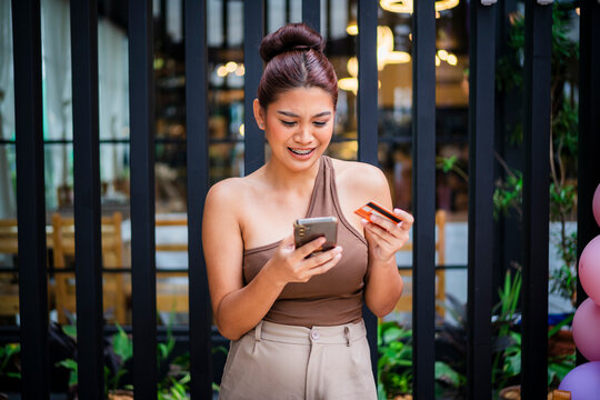 A stunned young Asian woman paying online with her phone and card in hand, visibly shocked by what she sees on screen while standing at a cafe.
