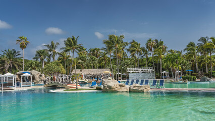 A relaxation area by the turquoise swimming pool. Deck chairs, awnings, arched pedestrian bridges with rope railings and lifebuoys. A pavilion with a thatched roof among palm trees. Blue sky, clouds.