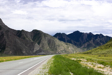 Long empty highway cuts through the plains of Altai, Russia, toward majestic mountain peaks. Overcast skies add drama to this wild and remote natural view. Road trip concept, copy space.