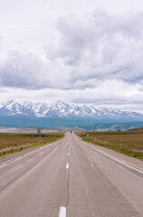Long empty highway cuts through the plains of Altai, Russia, toward majestic mountain peaks. Overcast skies add drama to this wild and remote natural view. Road trip concept, copy space.