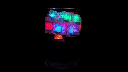 Bright LED ice cubes glowing inside the glass on a black background create colorful reflections and festive neon aesthetics of the drink. - Powered by Adobe