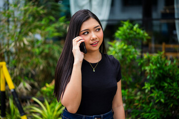A busy young Asian woman holding a phone to her ear while looking to the side, fully engaged in a focused conversation during a call in a garden