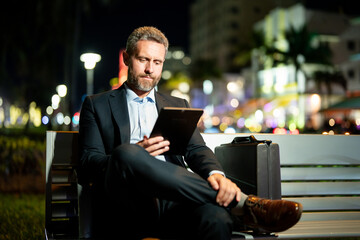 Online business. Mature business man working on tablet outdoors. Business man with tablet outdoor. Businessman with tablet on night street. Businessman using tablet, sit on bench. Night business.
