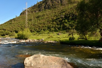 A huge stone in the bed of a beautiful mountain river and a power line on the bank on a sunny summer evening.