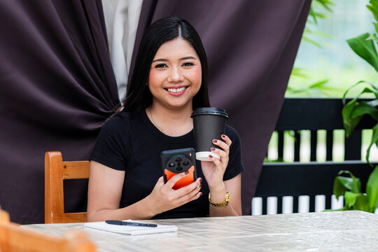 A cheerful young Asian woman multitasking with ease, smiling at the camera while using her phone and holding a coffee cup in her hand at a cafe. - Powered by Adobe