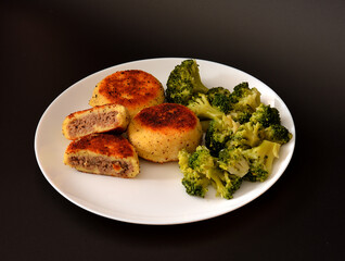 Potato cutlets stuffed with meat and boiled broccoli on a large round plate on a black background.