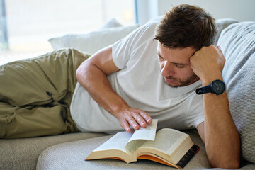 Obraz premium Close-up of man in casual clothes lying on sofa and reading book at home. Relaxation, leisure and self-education concept