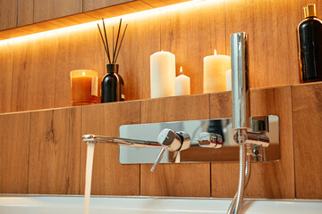 Modern chrome bathtub faucet pouring water into tub with foam and candles on shelf. Concept of home wellness, self care and relaxation in bathroom