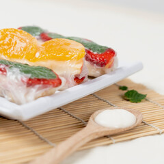 Three fresh fruit spring rolls with strawberries, orange slices, and mint leaves, neatly arranged on a white rectangular plate over a bamboo mat, accompanied by a wooden spoon with cream.