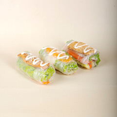 Three fresh spring rolls with various fillings grilled chicken, shrimp, and tofu wrapped in rice paper with lettuce and sprinkled with sesame seeds, arranged on a light beige background.