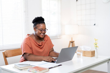Fototapeta premium Smiling African American woman working on laptop at home office, typing and looking at screen in bright modern workspace with books and documents on desk.