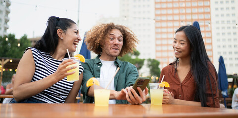 Three friends are laughing and enjoying refreshing drinks at an outdoor cafe, sharing a moment of connection and entertainment with a smartphone
