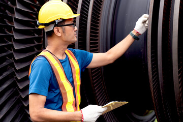 Closeup young Asian worker, power plant engineer in vest and helmet checking of a steam turbine's body, an essential component in electric power generation on outdoor with clipboard
