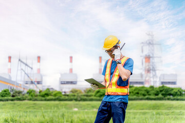 Image of young Asian power plant engineer working outdoors with tablet and radio communication on blurred of power plant and blue sky background. green energy power and environment concept
