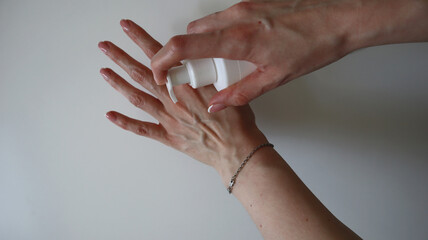 Photo of hand skin care. Hand holding a spray bottle over the other hand. Cream, sunscreen, soap, etc. White, thin hands, blood vessels visible. Thin chain bracelet on hand. White background.