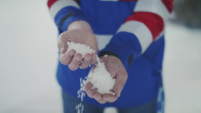 A medium close-up shot shows a person's hands pouring white, round pellets from one hand to another in slow motion. The product could be fertilizer, seeds, or de-icing salt.