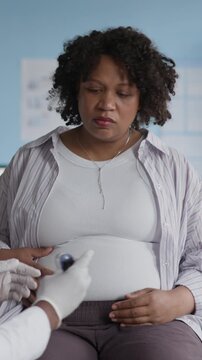 Vertical medium shot of Black male healthcare provider checking pregnant woman blood sugar level with glucometer, monitoring for gestational diabetes during routine prenatal checkup