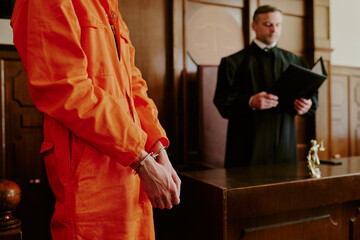 Caucasian young adult man in orange jumpsuit standing in handcuffs before judge in courtroom, judge...