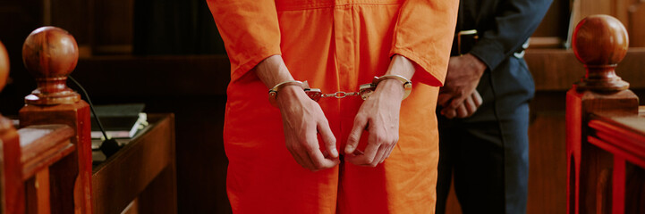 Caucasian young adult man standing in courtroom wearing handcuffs with hands in front, accompanied...