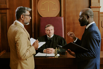 Caucasian middle aged male judge sitting behind bench while middle aged Black man and middle aged biracial man, standing in courtroom discussing legal case holding documents