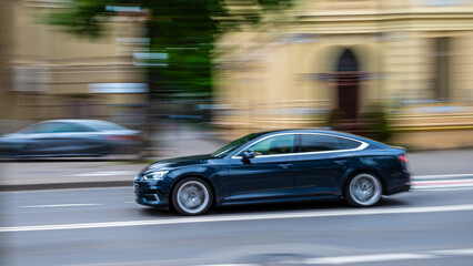 A black sedan is in motion, captured on a city street, creating a blurred background effect indicating speed.