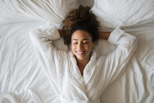 Young adult Black woman lying on bed with eyes closed, smiling and relaxing with hands behind head, wearing bathrobe, surrounded by bedding, enjoying peaceful morning